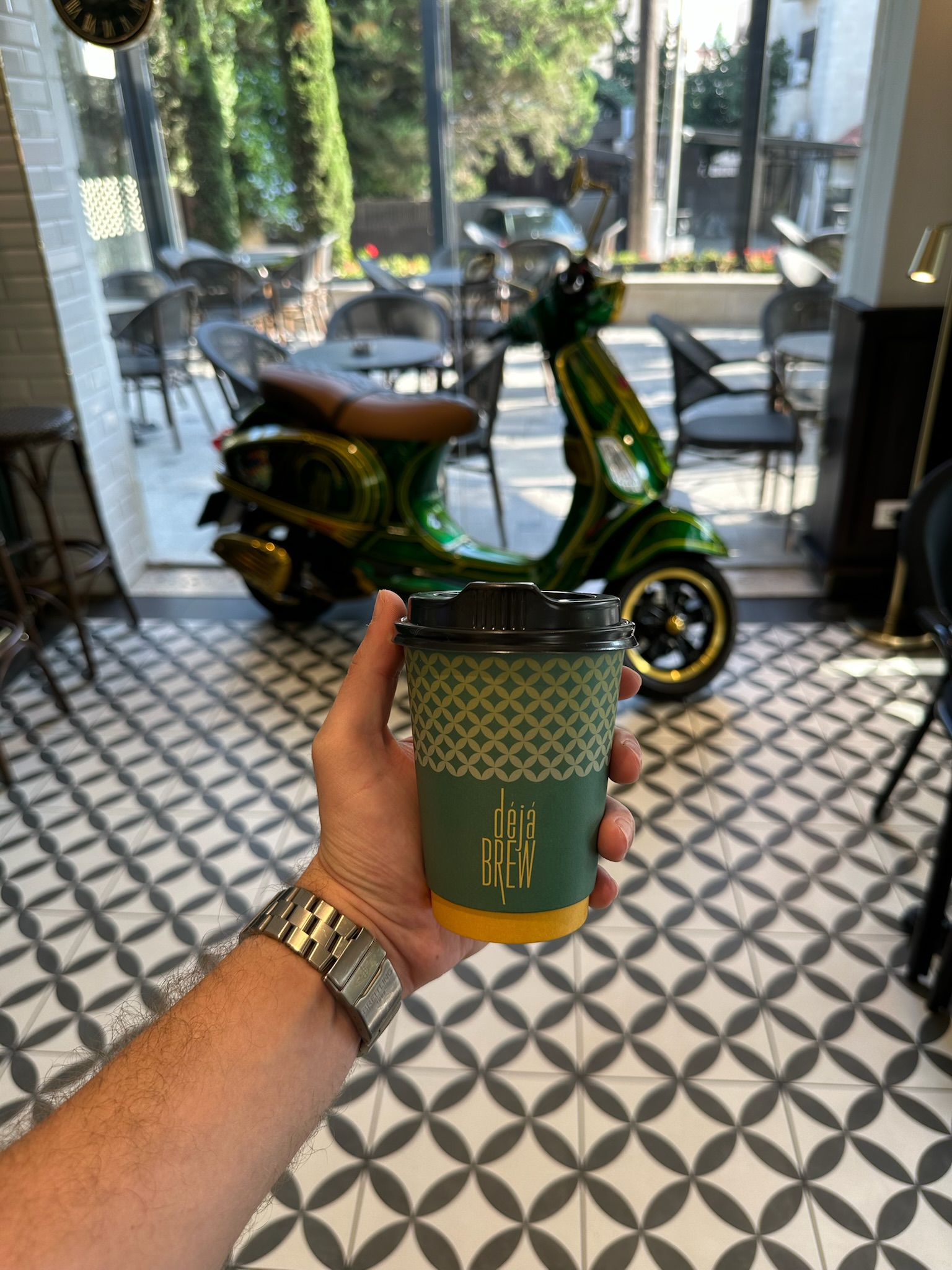 Deja Brew branded cup held inside the Abdoun café, green Vespa behind